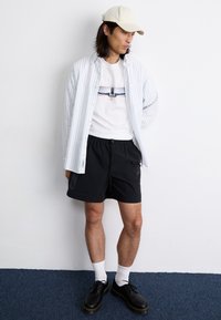 Model wearing a white t-shirt with a graphic, striped button-up shirt, black shorts, white socks, and black shoes with a yellow sole.