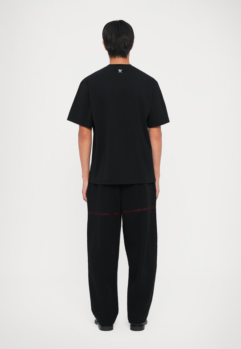 Sort kortærmet T-shirt med lige snit, med et hvidt logo på ryggen. Sammen med løstsiddende sorte bukser med røde stikninger.