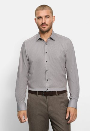 Man die een licht patroon overhemd met lange mouwen en knopen draagt, in een bruine broek gestopt met een donkere riem, staand tegen een effen achtergrond.