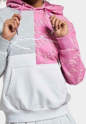 Personne portant un sweat à capuche avec un bloc de couleurs gris, rose à motifs et blanc, ajustant les cordons roses près du cou.