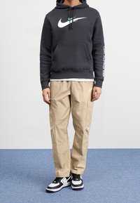Haut à capuche noir avec logo blanc de Nike et texte sur la manche, associé à un pantalon cargo beige et des baskets noir et blanc, sur une surface bleue.