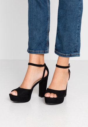 Fødder iført sorte ruskinds platform højhælede sko med ankelremme og cropped blå denim jeans på en lys gulvbagsgrund.