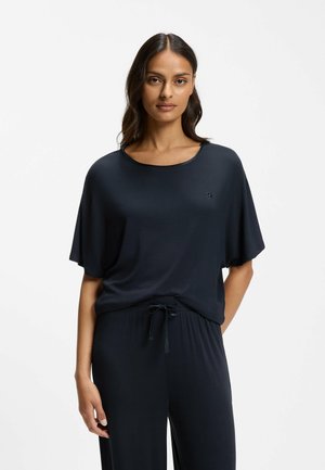 Donna in piedi contro uno sfondo semplice, indossa una maglia scura a maniche corte e pantaloni coordinati con coulisse, rivolta in avanti.