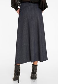 Frau in einem dunkel gestreiften Midikleid kombiniert mit schwarzen, hochhackigen Stiefeletten und einer schwarzen Bluse, die die Hände in den Taschen hat.