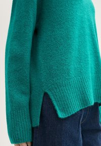 Türkiser Pullover mit strukturiertem Stoff, geripptem Saum und seitlichen Schlitzen, kombiniert mit dunklen Jeans. Das Design betont den Komfort.
