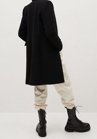 Henkilö, jolla on päällään musta polvipituinen takki, vaalea beige housut ja mustat nauhakenkäcombat-saappaat, seisoo kädet taskuissa.