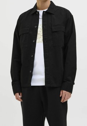 Person iført sort skjorte med knapper over hvid grafisk t-shirt og sorte bukser, stående mod en ensfarvet lys baggrund.