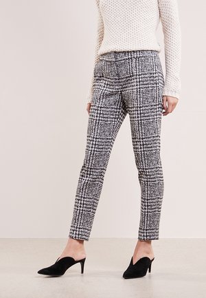Kobieta ubrana w dopasowane spodnie w czarno-białą kratę, biały długi sweter z fakturą oraz czarne mule na obcasie, stojąca na neutralnej podłodze.