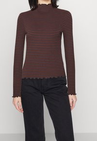 Långärmad, figurnära topp i mörkbrun med tunna vertikala ränder i ljusare nyanser. Har en vågig halsringning och nederkant, ribbad textur.