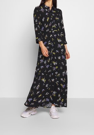Femme portant une robe maxi à boutons noire à motifs floraux, manches trois-quarts, et des baskets blanches, debout devant un fond uni.