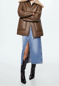 Giacca di pelle marrone con colletto in shearling, abbinata a una gonna di denim con spacco frontale e stivali neri di pelle al ginocchio.