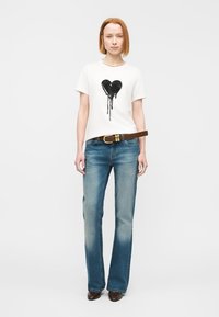 T-shirt blanc avec un motif de cœur noir, associé à un jean flare bleu clair. Ceinture tan avec des accessoires dorés et chaussures marron foncé.