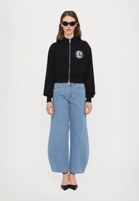 Melna zip-up sporta jaka ar apaļu logo uz krūtīm, apvienota ar plats kāju gaiši ziliem džinsiem un melniem norādītā pēdas augstpapēžu apaviem.