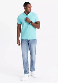 Camisa polo turquesa con cuello, combinada con jeans azul claro y zapatillas blancas. Reloj de pulsera notable en la muñeca izquierda.