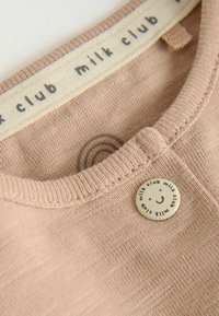 Gros plan sur le col d'un vêtement en maille beige avec une étiquette « milk club » et un bouton rond affichant un visage souriant et le texte « milk club ».
