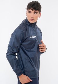 Giacca a vento blu Navy con collo alto, polsini elastici e tasche con zip. Presenta il logo "LEGEA" sul petto. Texture liscia.