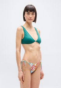 Γυναίκα με κοντά σκουρόχρωμα μαλλιά, φορώντας πράσινο τοπ μπικίνι και πολύχρωμο μπικίνι με δεσίματα στο πλάι, στέκεται μπροστά σε απλό ανοιχτό φόντο.