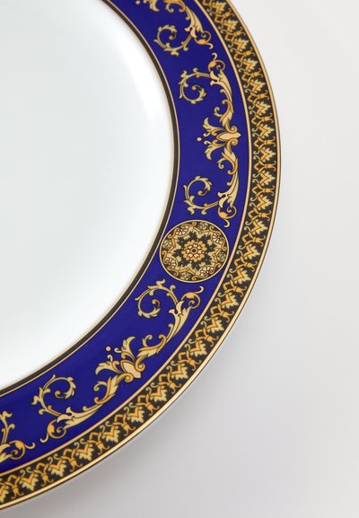 Balta porcelāna šķīvis ar zilu un zeltītu dekoratīvu apmali, kurā ir ziedu raksti un apaļi akcenti. Gluda tekstūra, elegants dizains.