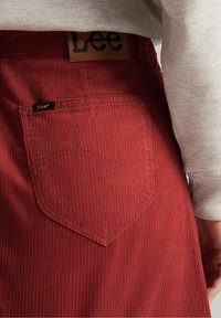 Pantalones de pana rojos con rayas verticales. Presenta un destacado bolsillo trasero con detalles de costura y una etiqueta de marca de cuero marrón.