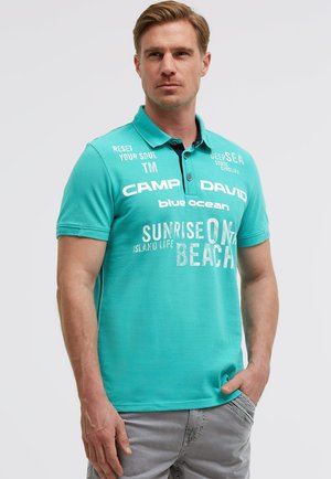 Mann trägt türkisfarbenes Poloshirt mit weißem aufgedrucktem Text, darunter „Camp David“ und „Sunrise on the Beach“, steht vor schlichtem Hintergrund.