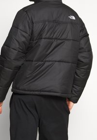 Chaqueta acolchada negra de nailon con un diseño acolchado, que presenta costuras horizontales y un logo en la parte superior de la espalda. Puños ajustables.