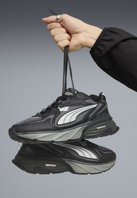 Czarne i szare trampki z siateczki i skóry, z białym paskiem jako akcentem i masywną podeszwą, trzymane w ręku.