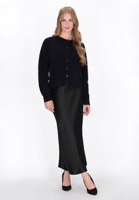 Cardigan lavorato a maglia blu navy con bottoni frontali, abbinato a una gonna nera in satin e scarpe con tacco nere, esposto su uno sfondo bianco.