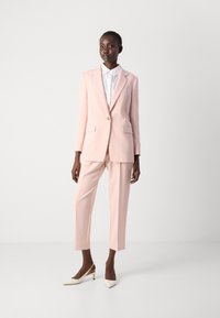 Hellrosa Anzug mit taillierter Jacke und passender Hose, darunter ein weißes Hemd mit Kragen und beige spitze High Heels.