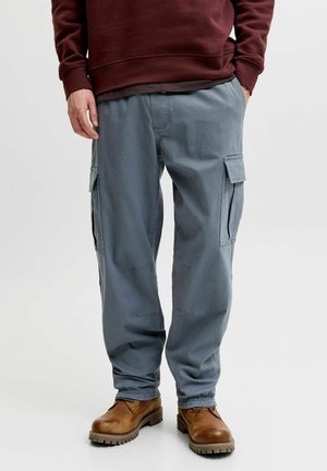Homme portant un pantalon cargo bleu avec des poignets élastiques et des bottes en cuir marron, debout avec une main dans la poche.