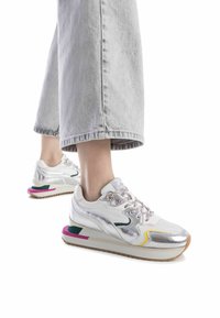 Zapatillas blancas con acentos metálicos, suela multicolor, cordones texturizados y una mediasuela acolchada. Llevadas con unos pantalones de mezclilla grises de pierna ancha.