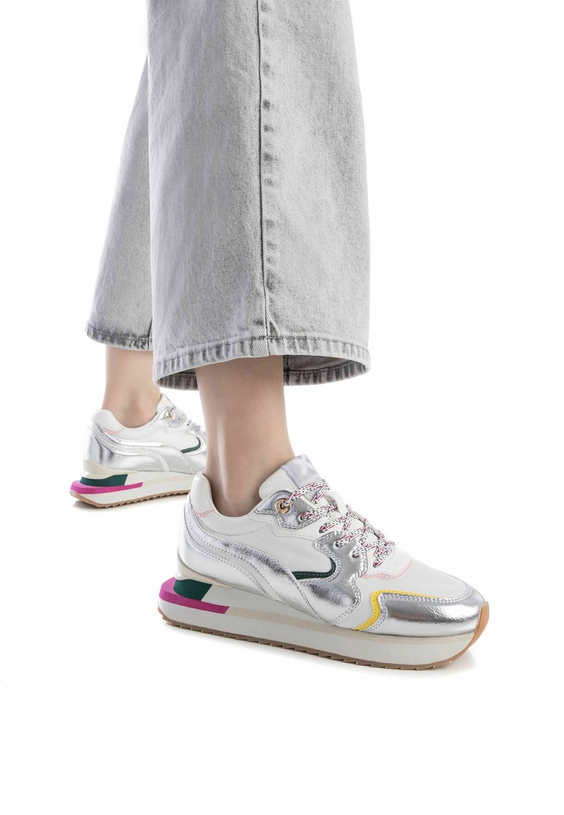 Zapatillas blancas con acentos metálicos, suela multicolor, cordones texturizados y una mediasuela acolchada. Llevadas con unos pantalones de mezclilla grises de pierna ancha.