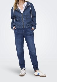 Jeansová bunda s kapucí, dvěma předními kapsami a elastickými manžetami; spárována s volnými džínovými kalhotami a mnohobarevnými teniskami.