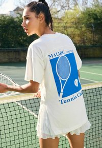 Vitt oversized t-shirt med blå grafik som visar en tennisklubba och texten "Malibu Tennis Club", i kombination med en veckad vit kjol.