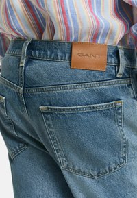 Blaue Denim-Jeans mit strukturierter Oberfläche, ausgestattet mit einem Leder-GANT-Logo-Patch am hinteren Bund und zwei Gesäßtaschen.