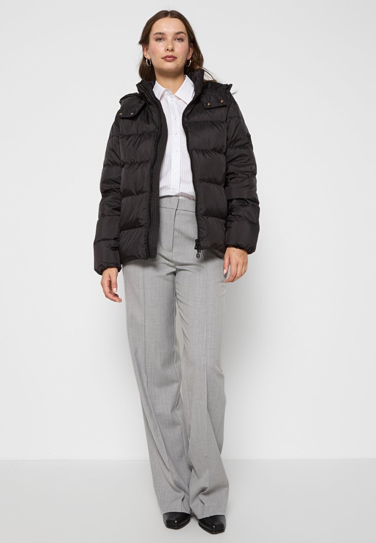 Frau in schwarzer Steppjacke, weißem Hemd und grauen weit geschnittenen Hosen, die vor einem schlichten weißen Hintergrund steht.