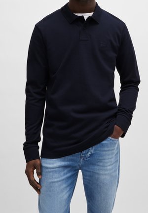 Mann trägt dunkelblaues Polohemd mit langen Ärmeln und geknöpftem Kragen sowie hellblaue Jeans, eine Hand in der Tasche vor einfarbigem Hintergrund.