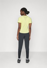 Helles gelbes, kurzärmliges Sporthemd mit einer figurbetonten Passform, kombiniert mit dunklen Slim-Fit-Hosen und schwarzen Laufschuhen. Reflektierende Akzente vorhanden.