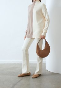 Personne portant un pull color block beige et crème, un pantalon blanc, des ballerines beige clair, tenant un sac en cuir marron en forme de croissant.