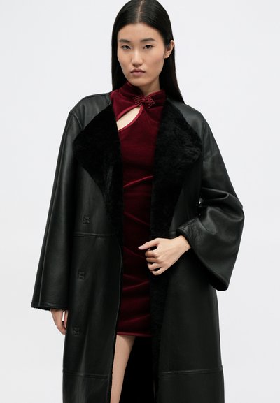 Casaco de pele preto com um colarinho peludo preto, apresentando um corte solto e mangas em sino. Por baixo, um vestido de veludo burgundy com um detalhe em forma de buraco.