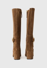 Botas de camurça castanhas até ao joelho com um salto quadrado, apresentando um fecho de correr lateral e detalhes em metal dourado perto do tornozelo.