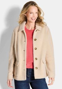 Beige fuzzy jasje met een kraag, vijf bruine knopen en twee zijzakken, gelaagd over een koraalkleurig shirt en blauwe spijkerbroek.