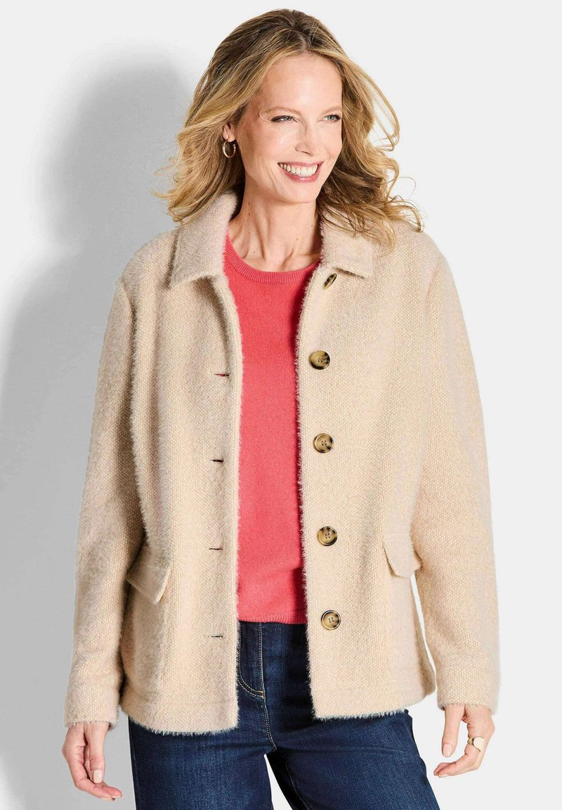 Beige fuzzy jasje met een kraag, vijf bruine knopen en twee zijzakken, gelaagd over een koraalkleurig shirt en blauwe spijkerbroek.