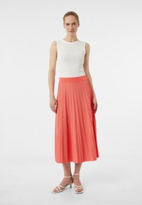 Biała top bez rękawów z prążkowanej tkaniny i koralowa, plisowana spódnica maxi z pionowymi paskami, w połączeniu z białymi sandałkami na obcasie.