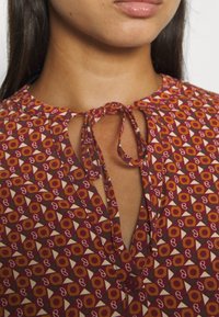 Bordeauxfarbene Bluse mit geometrischem Print, ausgestattet mit einem Schlüsselloch-Ausschnitt und einem Bindedetail. Der Stoff hat eine glatte Textur und einen dezenten Glanz.