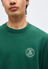 Νεαρό άτομο που φοράει πράσινο φούτερ με στρογγυλό λευκό λογότυπο που γράφει "FC Marc O'Polo Stockholm 1967" στο αριστερό στήθος.