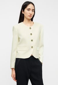 Κρεμ σακάκι με στρογγυλή λαιμόκοψη, πέντε κουμπιά με χρυσές λεπτομέρειες και τσέπες μπροστά, σε συνδυασμό με μαύρο παντελόνι.