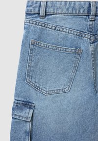 Tessuto di denim azzurro chiaro, con una tasca posteriore a doppia cucitura dal design angolato. Include un piccolo dettaglio a patta sul lato.