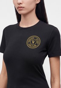 Γυναίκα φοράει μαύρο κοντομάνικο T-shirt με κίτρινο κυκλικό λογότυπο "Versace Jeans Couture" στο αριστερό στήθος.