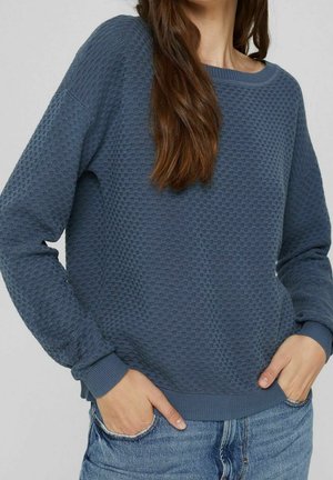 Frau trägt einen strukturierten blauen Langarm-Pullover mit gerippten Bündchen und Saum, kombiniert mit hellblauen Jeans, die Hände in den Taschen.