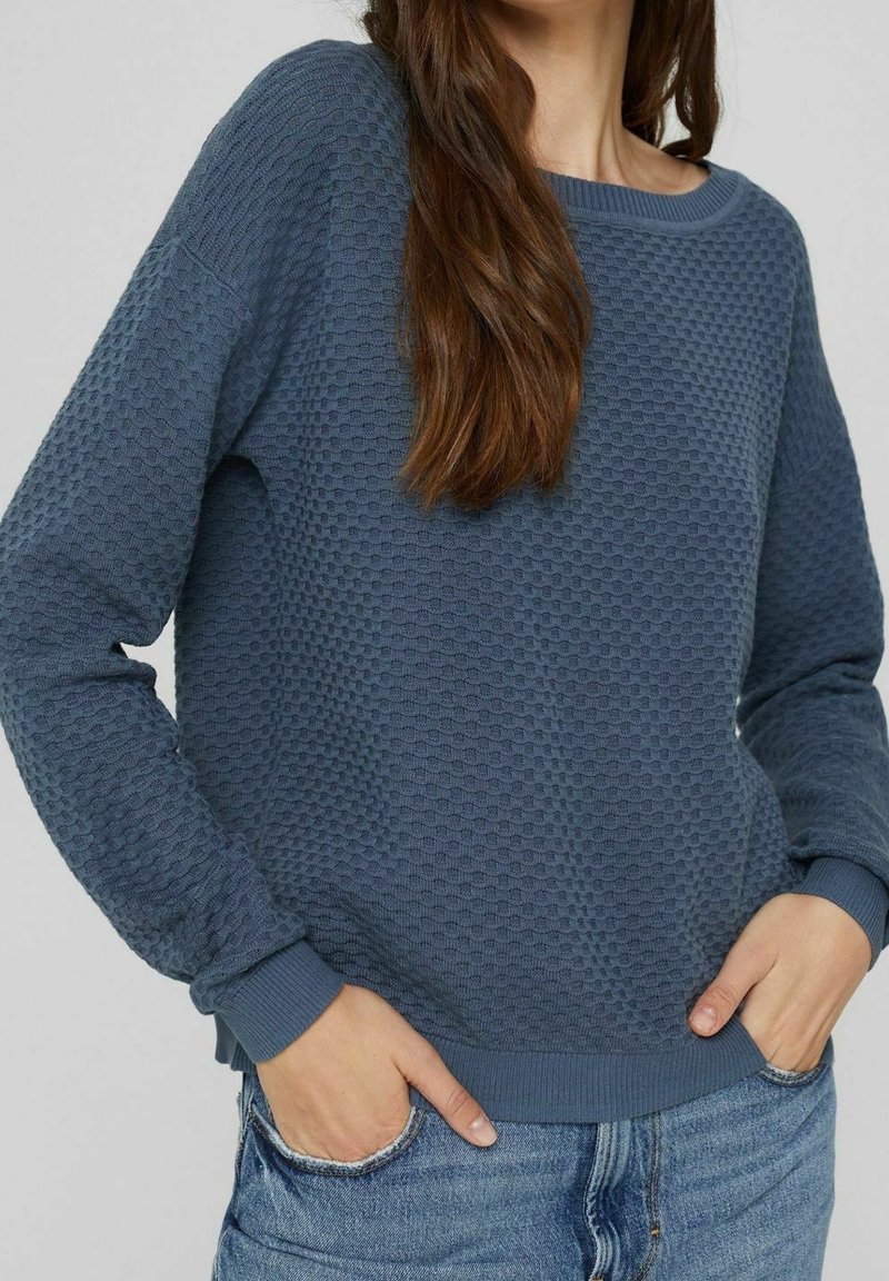 Femme portant un pull bleu texturé à manches longues avec des poignets et un ourlet côtelés, associé à un jean en denim bleu clair, les mains dans les poches.